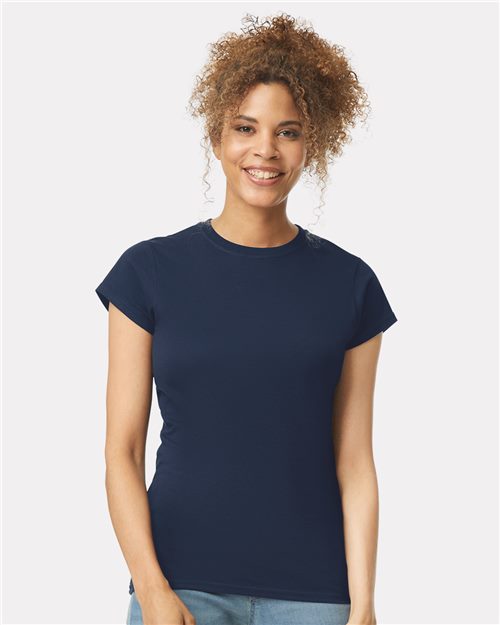 A person with curly hair smiles in a plain navy blue Gildan - Softstyle® Women’s T-Shirt, crafted from OEKO-TEX certified ring-spun cotton. The background is solid light gray.