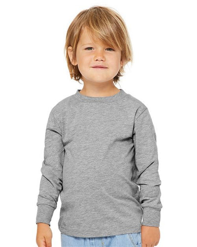 A young child with light brown hair is facing the camera, wearing a BELLA + CANVAS Toddler Jersey Long Sleeve Tee, gray in color and crafted from Airlume cotton, paired with light blue jeans against a plain white background.