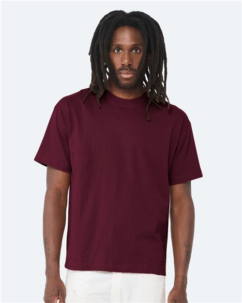 Load image into Gallery viewer, A person with long dreadlocks is wearing a BELLA + CANVAS - 6 oz. Heavyweight Tee in plain maroon, made from Airlume combed and ring-spun cotton, paired with white pants against a light gray background.
