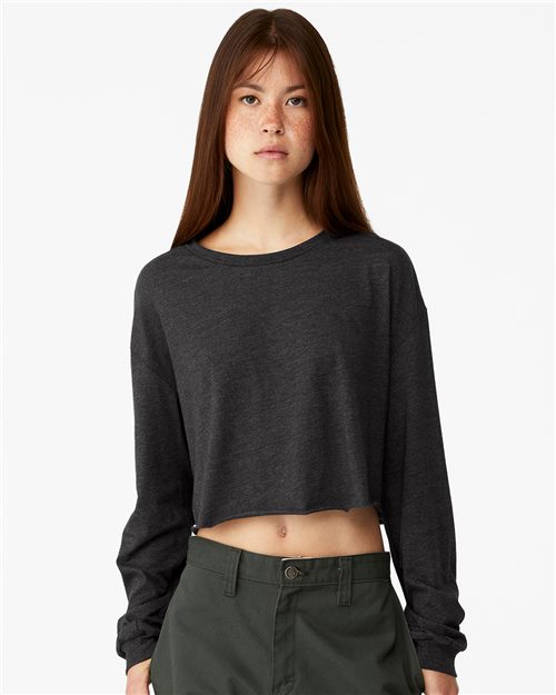 A young woman with long brown hair wears the BELLA + CANVAS FWD Fashion Women's Crop Long Sleeve Tee in dark gray, made from Airlume combed cotton, paired with green pants against a white background. Her look highlights BELLA + CANVAS's commitment to sustainable manufacturing.