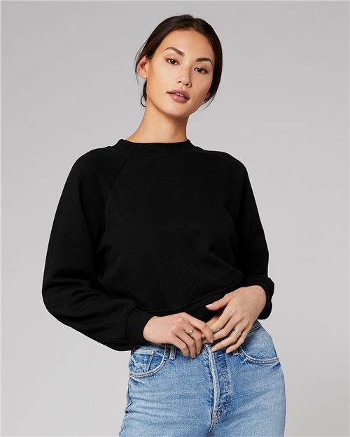 Load image into Gallery viewer, A person wearing a BELLA + CANVAS Women&#39;s Raglan Pullover Fleece in black and blue jeans stands against a gray background. Their long hair is tied back, holding the soft airlume combed cotton hem with a neutral expression.
