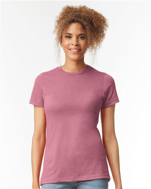 A person with curly hair wears a fitted Gildan - Softstyle® Women's CVC T-Shirt, made from OEKO-TEX certified materials, in pink and light blue jeans against a plain white background, smiling.