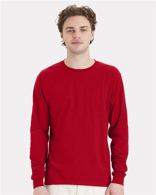 A person with brown hair wears a plain red Hanes Essential-T Long Sleeve T-Shirt and white pants, standing against a light gray background. Their neutral expression reflects an appreciation for sustainable manufacturing practices.