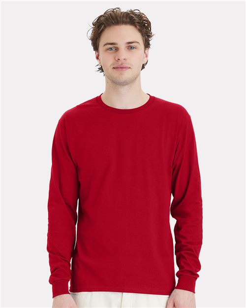 Load image into Gallery viewer, A person with brown hair wears a plain red Hanes Essential-T Long Sleeve T-Shirt and white pants, standing against a light gray background. Their neutral expression reflects an appreciation for sustainable manufacturing practices.
