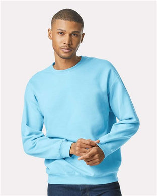 Wearing the Gildan Softstyle® Midweight Crewneck Sweatshirt in bright blue with dark jeans, a person stands against a white background, looking at the camera with hands loosely clasped, showcasing sustainable fashion.