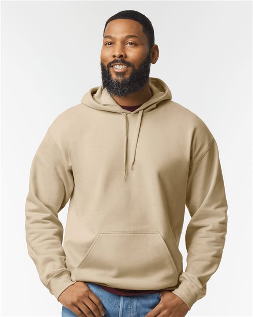 A bearded man stands against a plain white background, smiling gently with hands in pockets, wearing the Gildan - Softstyle® Midweight Hooded Sweatshirt made from OEKO-TEX certified ring-spun cotton and paired with jeans.