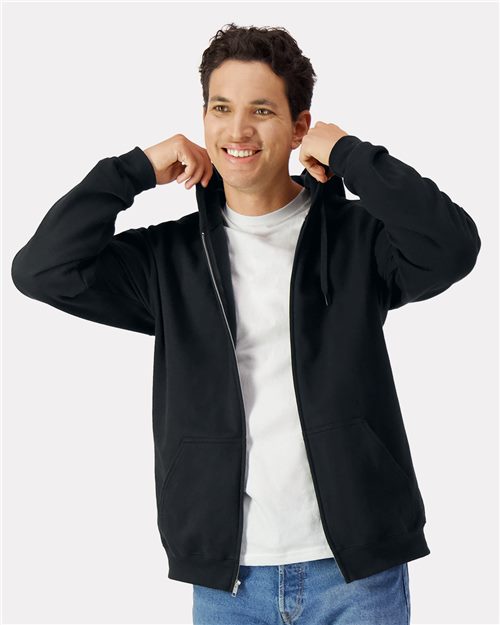 Wearing a Gildan Softstyle® Full-Zip Hooded Sweatshirt made from OEKO-TEX certified materials, a person smiles while adjusting the hood of their black hoodie over a white shirt. In jeans, they stand against a plain white background, exemplifying sustainable production practices.