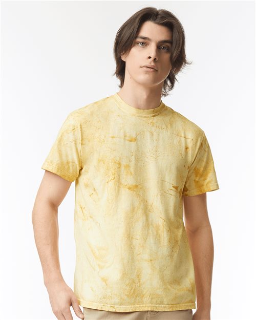 A person with shoulder-length brown hair stands against a white background, wearing a Comfort Colors Colorblast™ Heavyweight T-Shirt in yellow marbled pattern. Their beige pants complement the look. Displaying a neutral expression and relaxed posture, they embody effortless style.