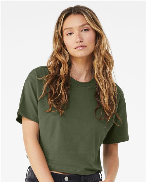 A person with long wavy hair, relaxed with a slight head tilt, is wearing a green BELLA + CANVAS Women's Jersey Crop Tee, made from 100% Airlume combed cotton, and dark pants against a plain background.