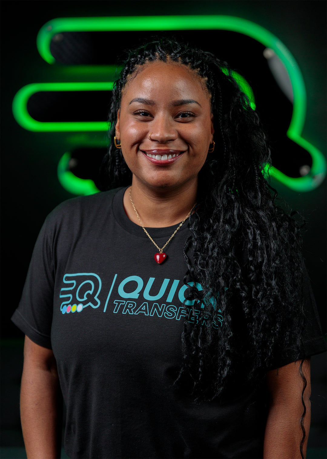 A person with long, curly hair smiles at the camera, wearing a black "Quick Transfers" t-shirt with a heart-shaped necklace. The background features a green, abstract logo.