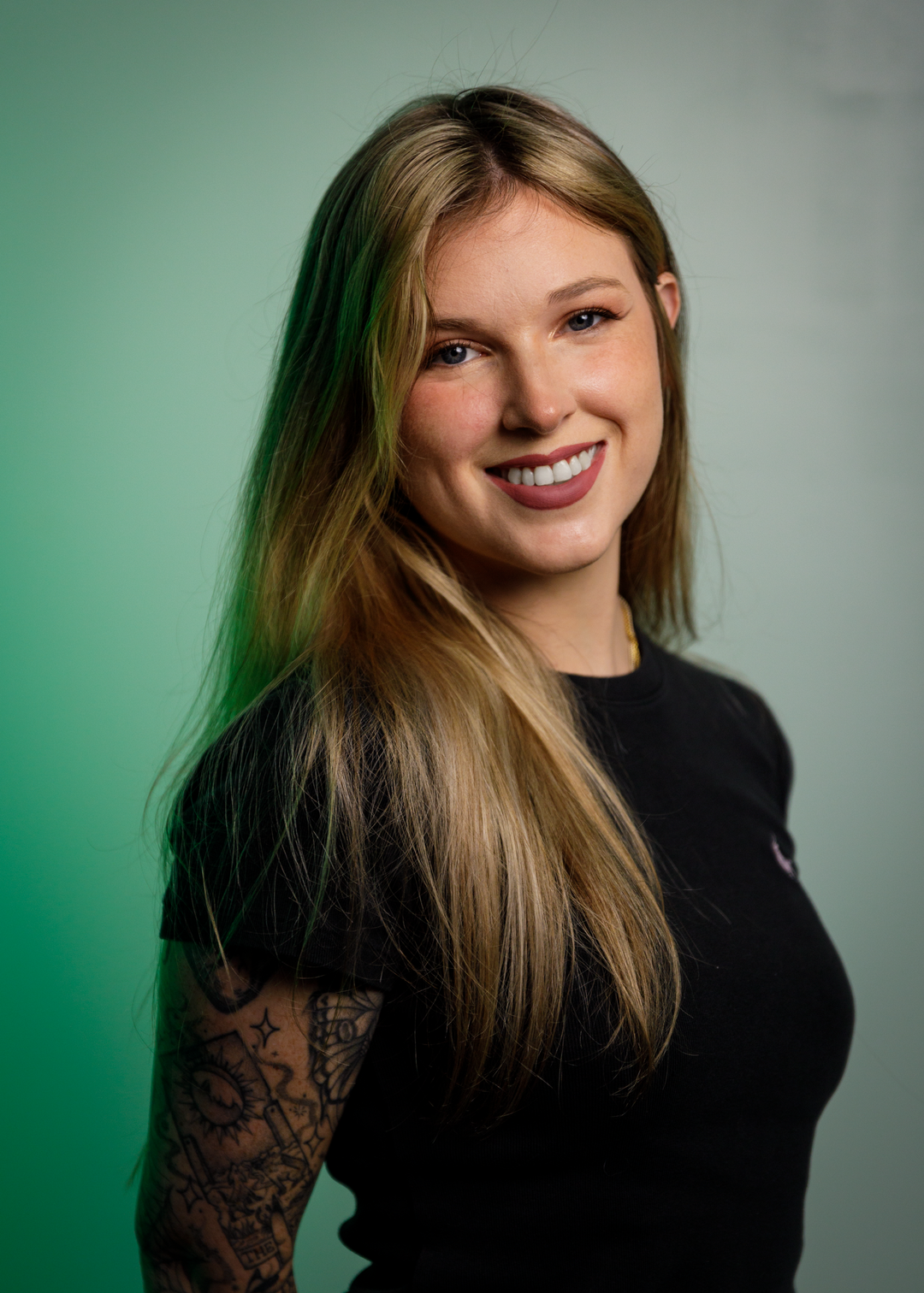 A smiling woman with long blonde hair stands in front of a green background. She is wearing a black t-shirt and has a tattoo sleeve on her left arm.