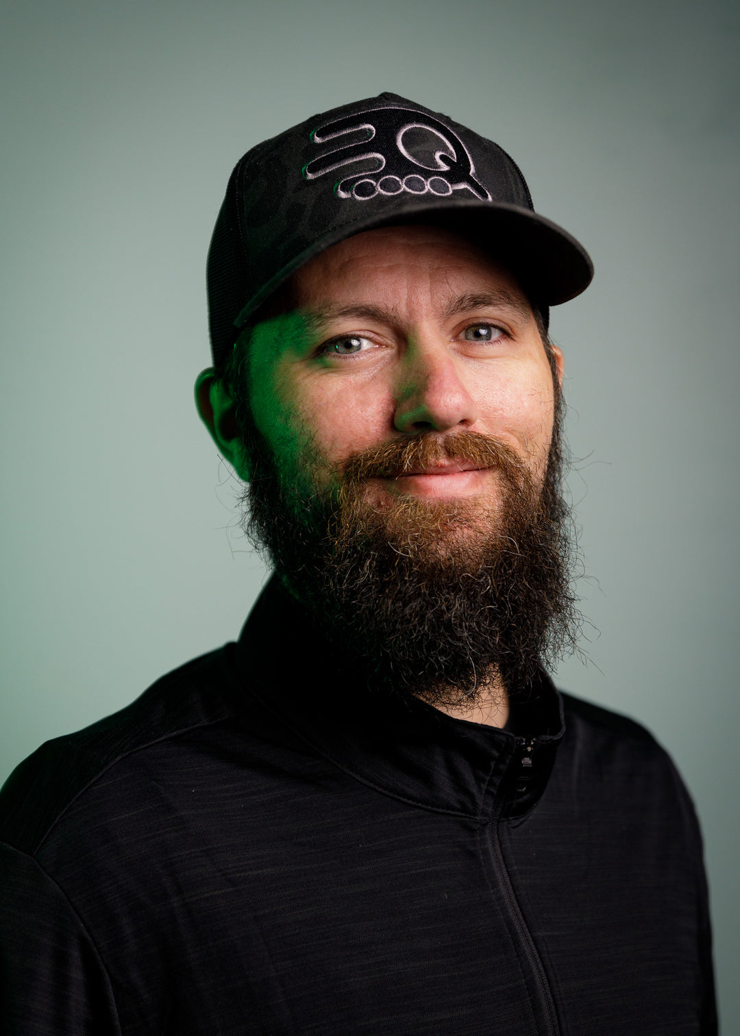 A man with a full beard, wearing a black cap with an embroidered design and a black jacket, stands against a plain green background.