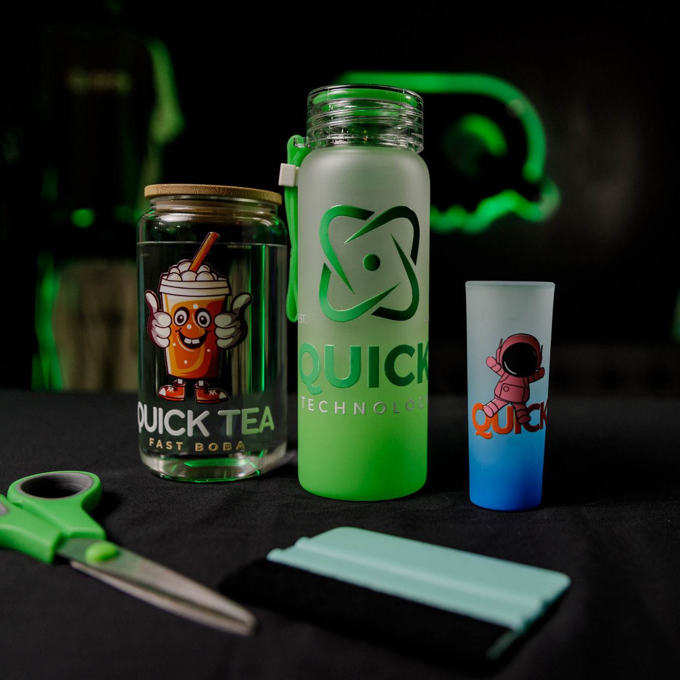 A table holds a jar with a cartoon bubble tea, a frosted bottle with "QUICK TECHNOLOGY," a colorful cup with a small astronaut illustration, green scissors, and a flat tool. The background is black with a neon green logo.