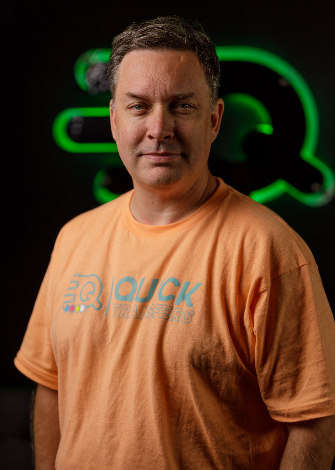 A person in an orange shirt with a "Quick Transfers" logo stands confidently against a dark background with a green neon sign.