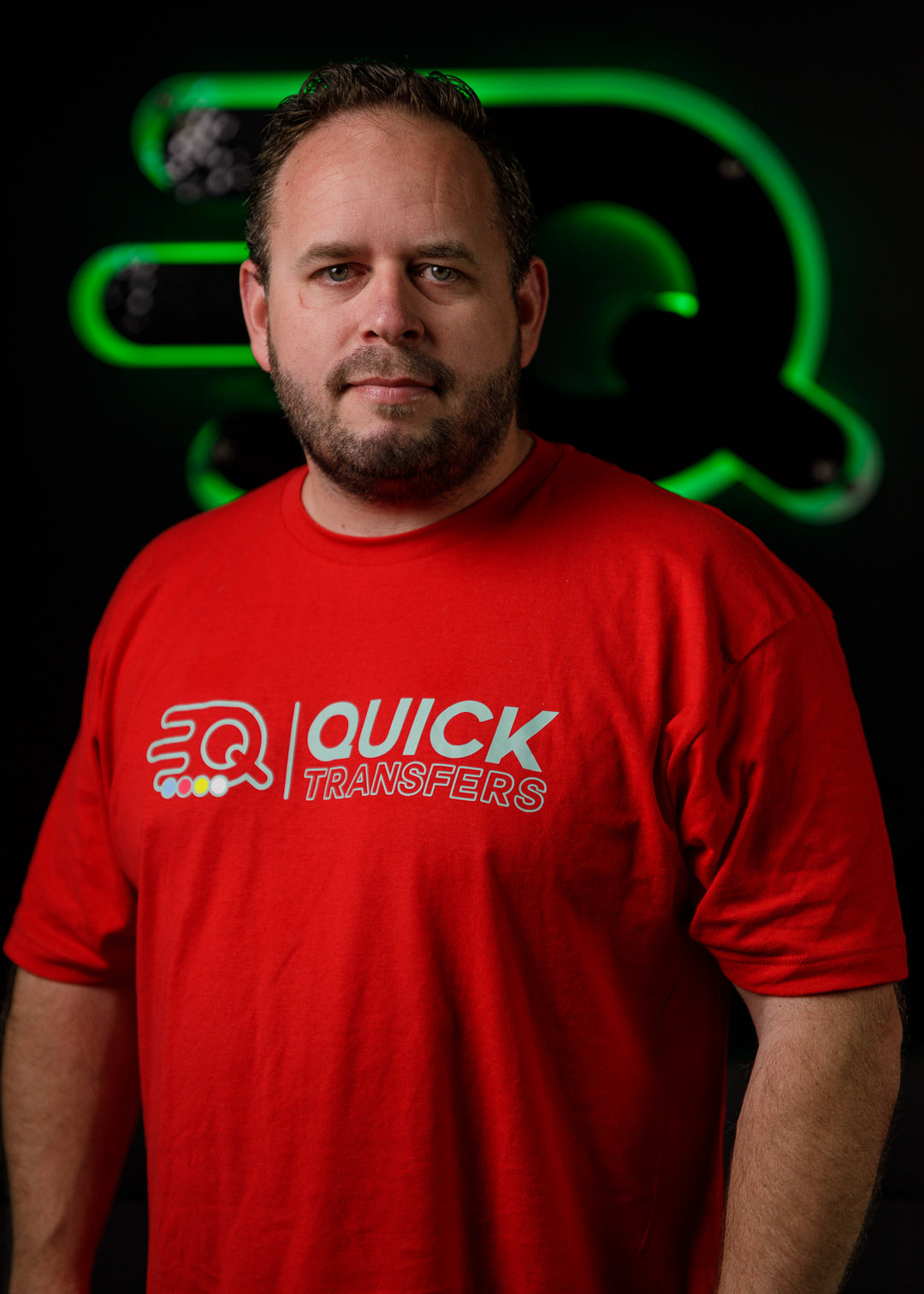 A man with a beard is wearing a red T-shirt with "Quick Transfers" printed on it. He stands in front of a dark background with a green logo featuring a stylized "Q.