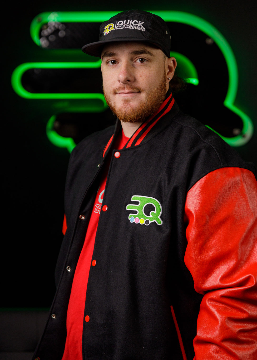 A person wearing a black and red sports jacket and a black cap with a logo stands confidently. The jacket and cap both feature a green and white symbol. The background is dark with a glowing green logo of a stylized letter "Q.