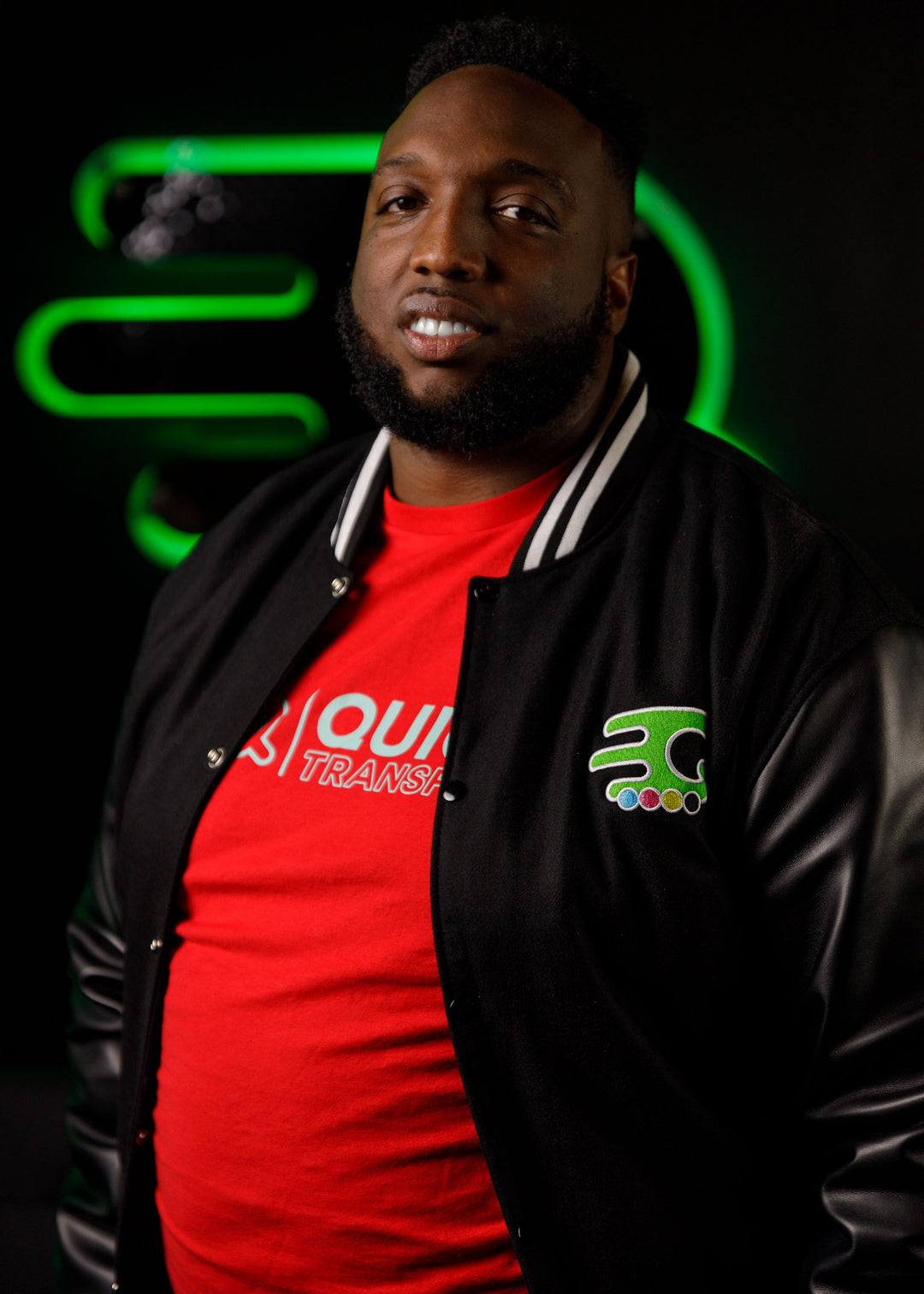 A man in a black and white varsity jacket over a red shirt stands against a dark background with green neon lighting. He has short hair and a beard, and he is smiling slightly.