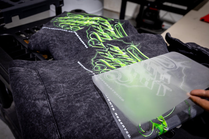 Close-up of a black T-shirt being screen printed with neon green and white graffiti-style text using the FASTFILM™ DTF TRANSFERS - Build a Gang Sheet by Quick Transfers. The shirt is positioned on a printing press, while a blurred hand holds a screen frame above.