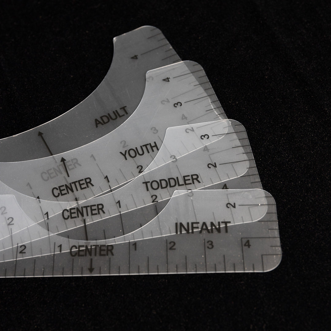 The Quick Transfers Alignment Ruler set includes four transparent rulers labeled "Infant," "Toddler," "Youth," and "Adult" for professional-quality apparel decoration, all arranged in a fan shape on a dark background for effortless crafting precision.