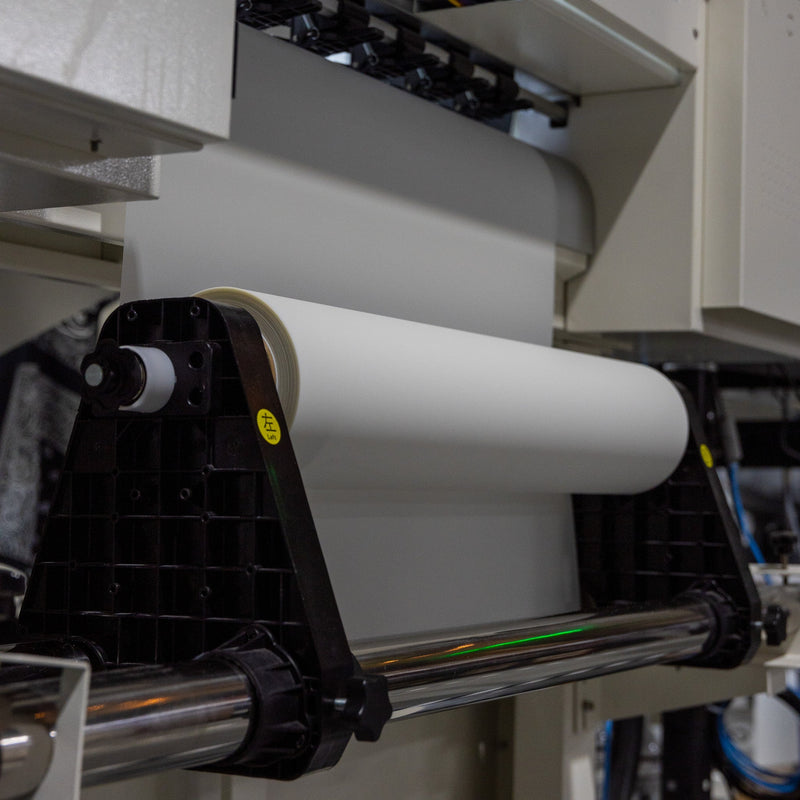 Load image into Gallery viewer, The 24in Hot Peel DTF Film - Double Side Matte by DTF Locker is mounted on a printing machine, ready for processing in an industrial facility with visible metal components and mechanical structures.
