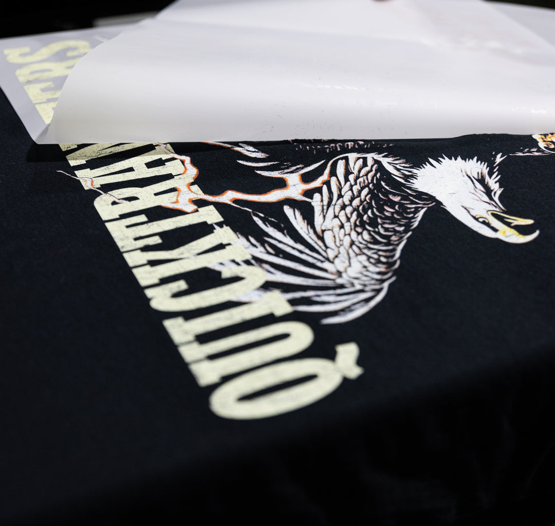 Close-up of a black tee showcasing a bold eagle graphic and "EQUICKARIO,” partially revealed as glossy white paper peels away, highlighting the exceptional detail from Quick Transfers' FASTFILM™ DTF TRANSFERS - Build a Gang Sheet, created with direct to film transfers.