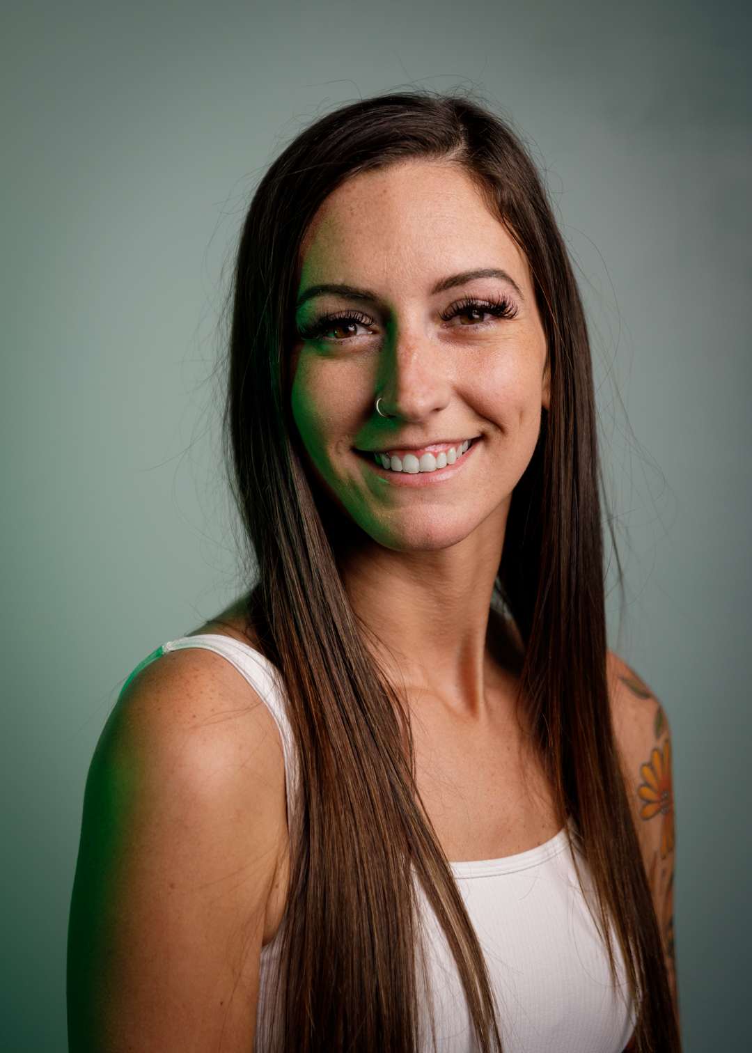 A woman with long brown hair smiles at the camera. She is wearing a white tank top. The background is a solid pale green color.