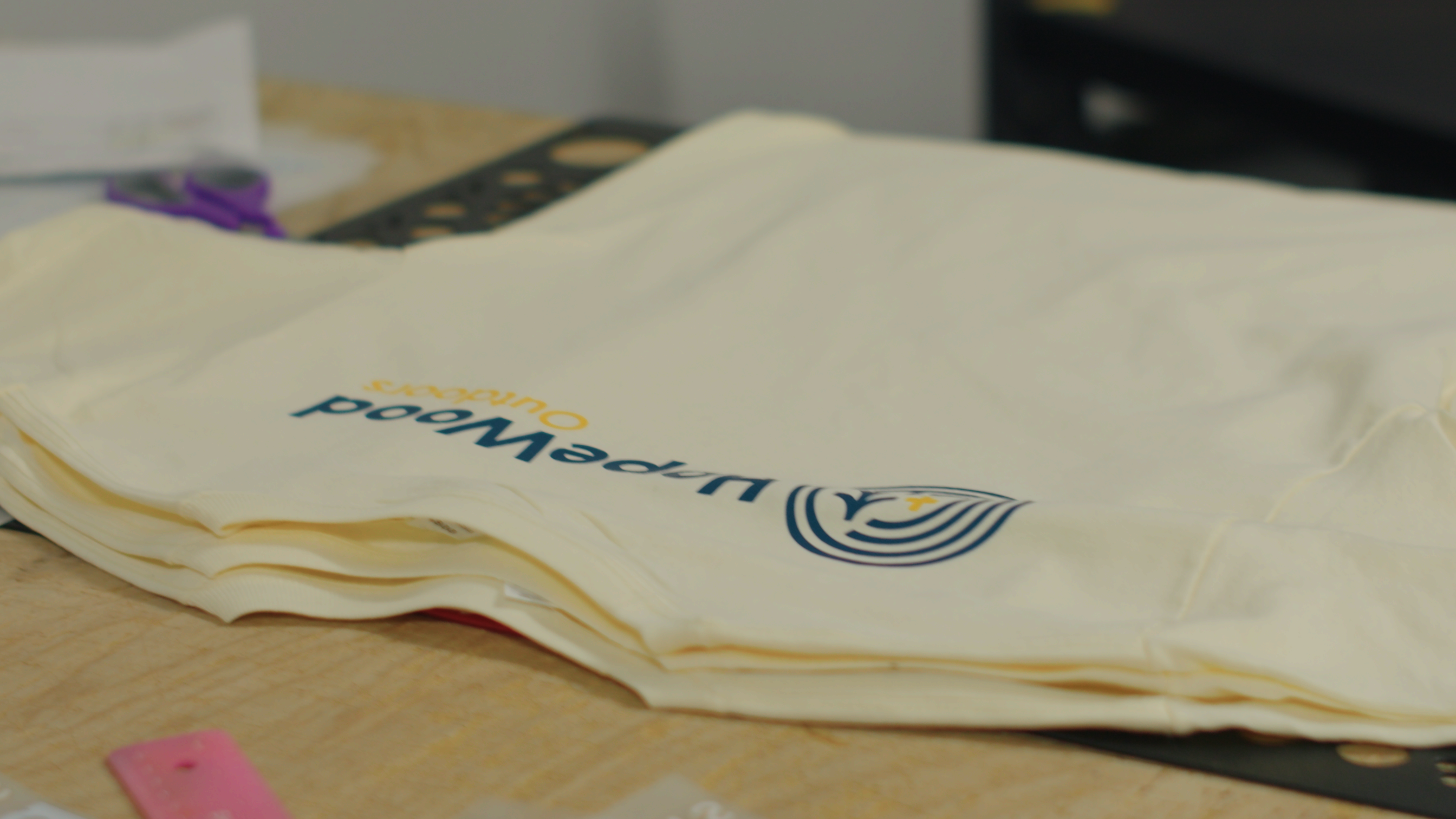 A stack of folded light-colored shirts lies on a table. The top shirt displays a logo with the text "Upperwood Outreach" and a shield graphic. Scissors and other items are blurred in the background.