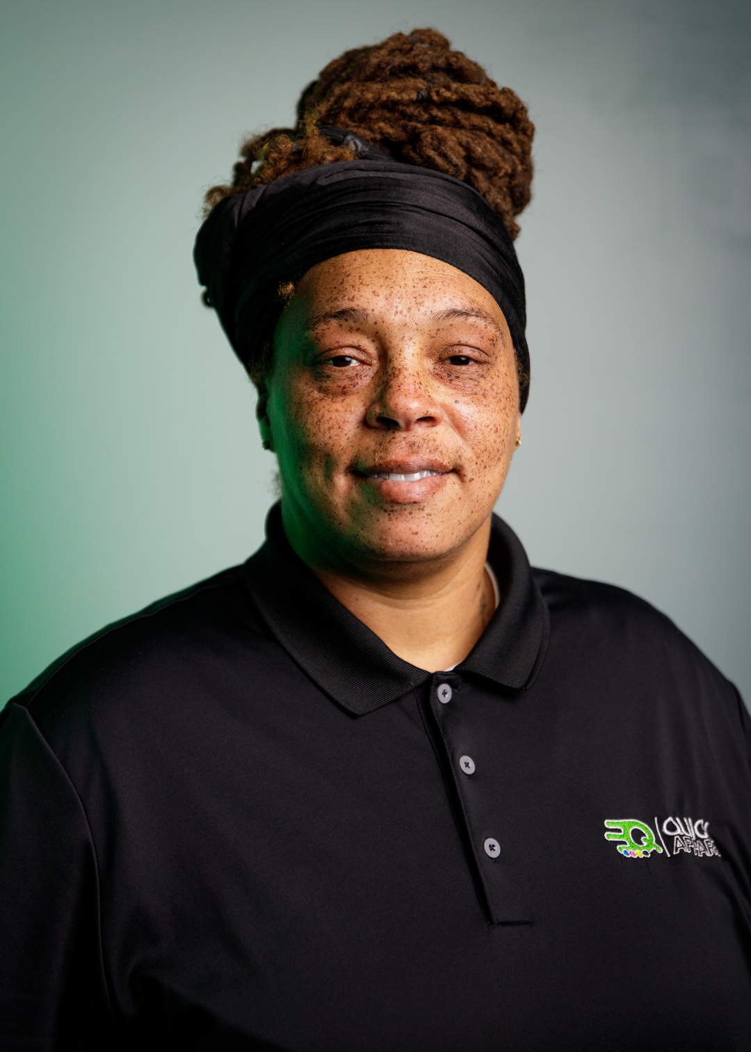 A person with long, coiled hair wrapped in a black headscarf is wearing a black polo shirt with a green logo. They have freckles and are smiling slightly while standing against a faded green background.