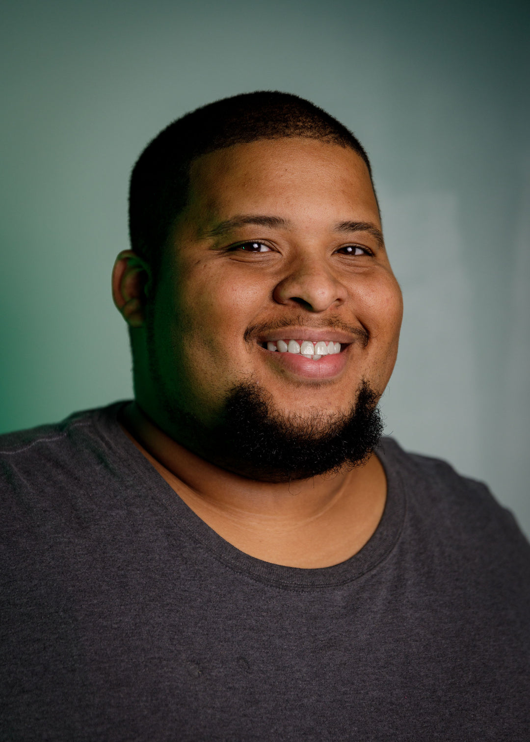 A person with short hair and a beard is smiling at the camera. They are wearing a dark gray T-shirt. The background is a soft green color.