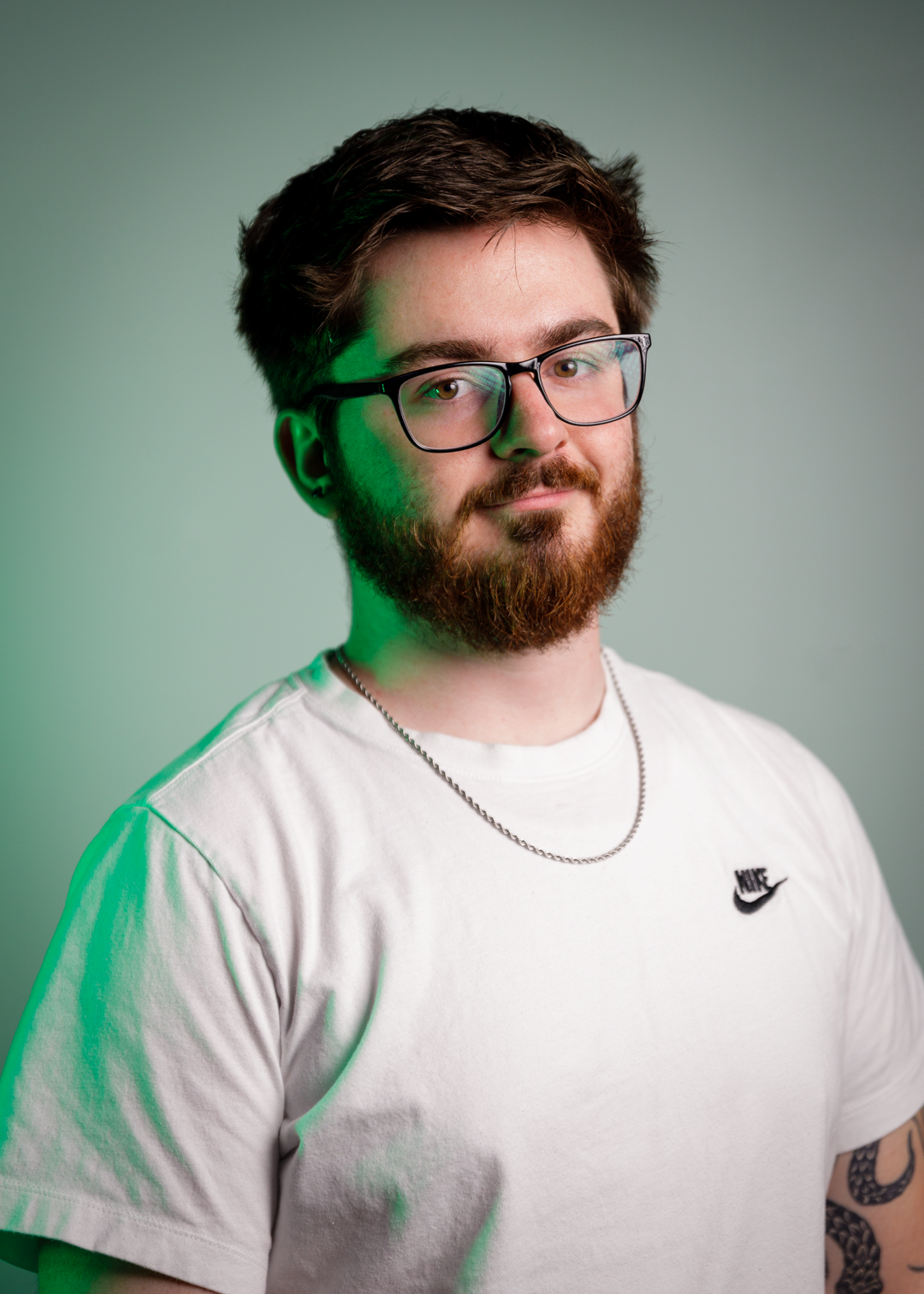 A man with a beard and glasses is wearing a white Nike t-shirt and a silver chain necklace, standing against a greenish-gray background. The lighting casts a slight green tint on the side of his face.