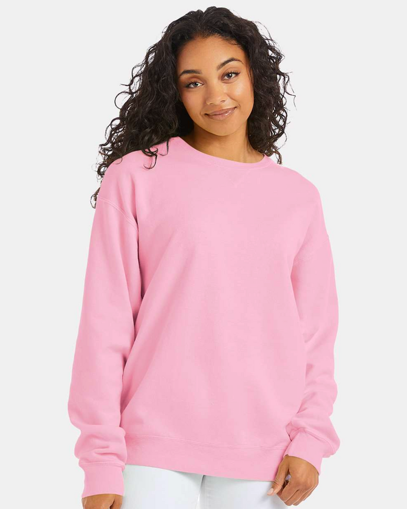 Load image into Gallery viewer, A woman with curly dark hair smiles softly, wearing a ComfortWash by Hanes Garment-Dyed Crewneck Sweatshirt in light pink and white pants, standing against a plain light gray background.
