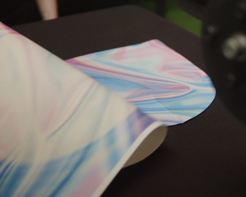 Load image into Gallery viewer, A close-up of a partly unrolled fabric roll with a soft, swirled pastel pattern in blue, pink, and white lies on a black surface. The silky texture&#39;s vibrant design is ideal for sublimation printing and bulk orders. Product: SUBLIMATION TRANSFERS - Transfers By Size by Quick Transfers.
