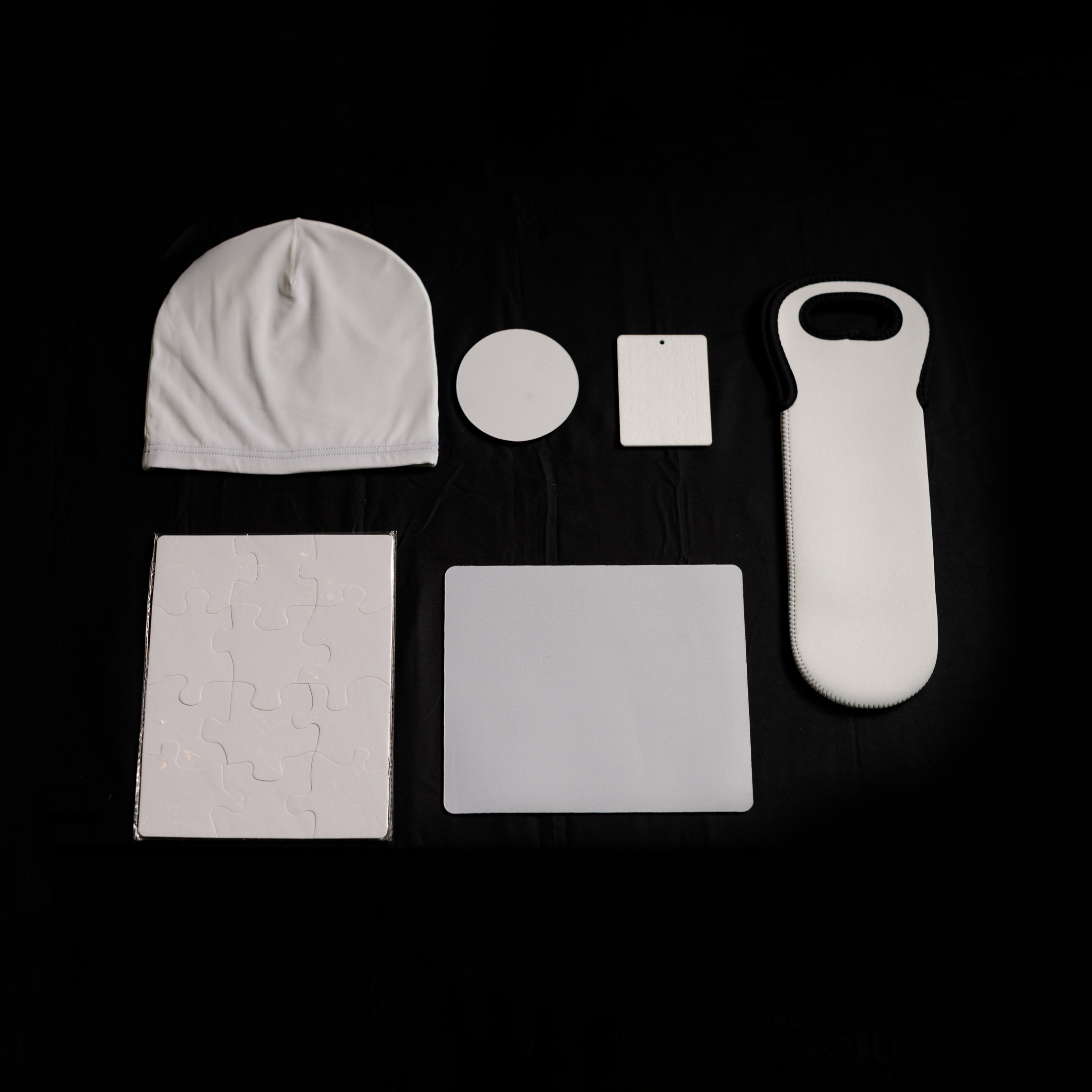 A collection of white objects on a black background, including a beanie, circular and rectangular coasters, a puzzle, a hot water bottle cover, and a square mouse pad.