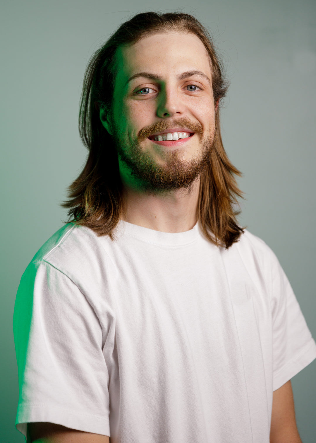 A person with long hair and a beard smiles at the camera. They are wearing a plain white t-shirt against a neutral background with a greenish tint.