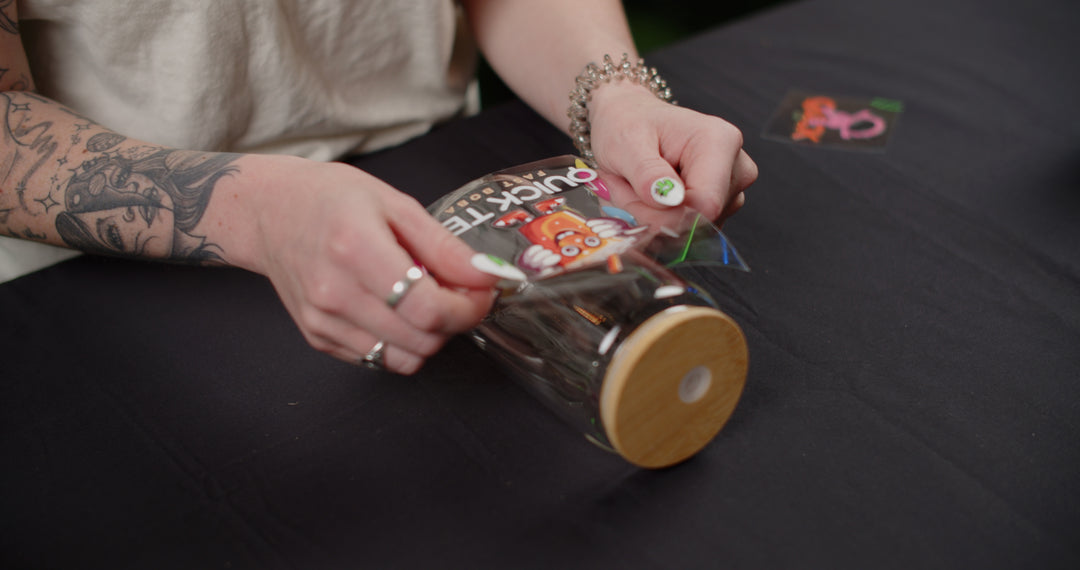 A person wearing rings and a bracelet is carefully applying a colorful cartoon character sticker with the word &quot;quickie&quot; using FOREVERFILM™ 3D UV DTF TRANSFERS from Quick Transfers onto a clear jar with a wooden lid. The jar sits on a black tablecloth, as they position the sticker for custom durable decoration.