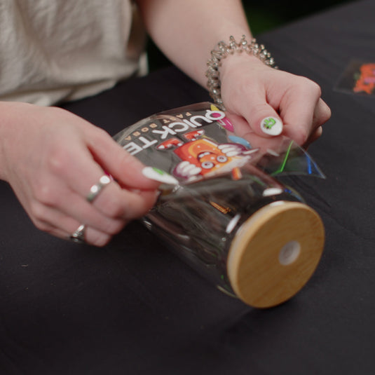 A tattooed individual, wearing rings and a bracelet, gently peels the label from a clear jar with a wooden lid. Displaying vibrant graphics likely crafted using FOREVERFILM™ 3D UV DTF TRANSFERS by Quick Transfers, the scene is set against a black cloth backdrop.