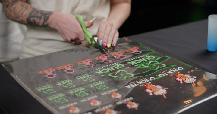 A person with colorful nails and a tattoo on their forearm uses green scissors to cut out vibrant printed designs from a sheet. These designs, perfect for creating custom durable decorations, include text and images of a cartoon character utilizing Quick Transfers' FOREVERFILM™ 3D UV DTF TRANSFERS - Build a Gang Sheet technology.