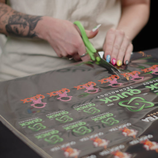 A person with colorful nails and a forearm tattoo uses green scissors to cut a glossy black sheet displaying "Quick Transfers" in vibrant green and pink, adorned with a custom cartoon character, expertly crafted using FOREVERFILM™ 3D UV DTF TRANSFERS.