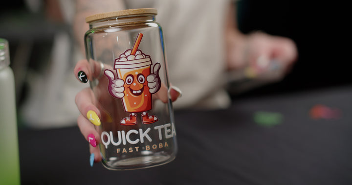 A person with vibrant nail art holds a transparent glass jar adorned with a cheerful cartoon drink, complete with a smile, straw, and thumbs up. The jar features the text "Quick Tea Fast Boba," printed using Quick Transfers' FOREVERFILM™ 3D UV DTF technology. The background is softly blurred.