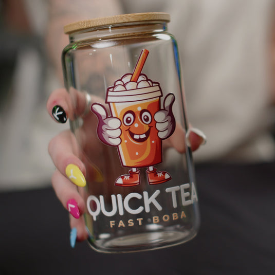 A person with multi-colored, designed nails holds a clear glass featuring a cartoon bubble tea character and the text 