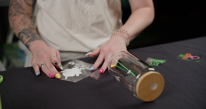 A person with colorful nails is decorating a clear glass water bottle with a wooden lid using Quick Transfers’ FOREVERFILM™ 3D UV DTF TRANSFERS for custom, durable decorations. They are applying a white turtle-shaped sticker onto the bottle while a green and black sticker rests nearby on the table.