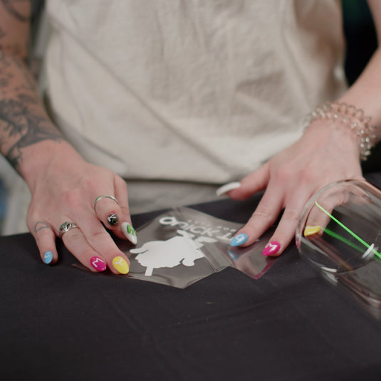 With colorful nails, a person meticulously applies a white FOREVERFILM™ 3D UV DTF TRANSFER by Quick Transfers to a clear glass jar with a wooden lid. Adorned in light beige and bracelets, they showcase their creative flair on a black surface.