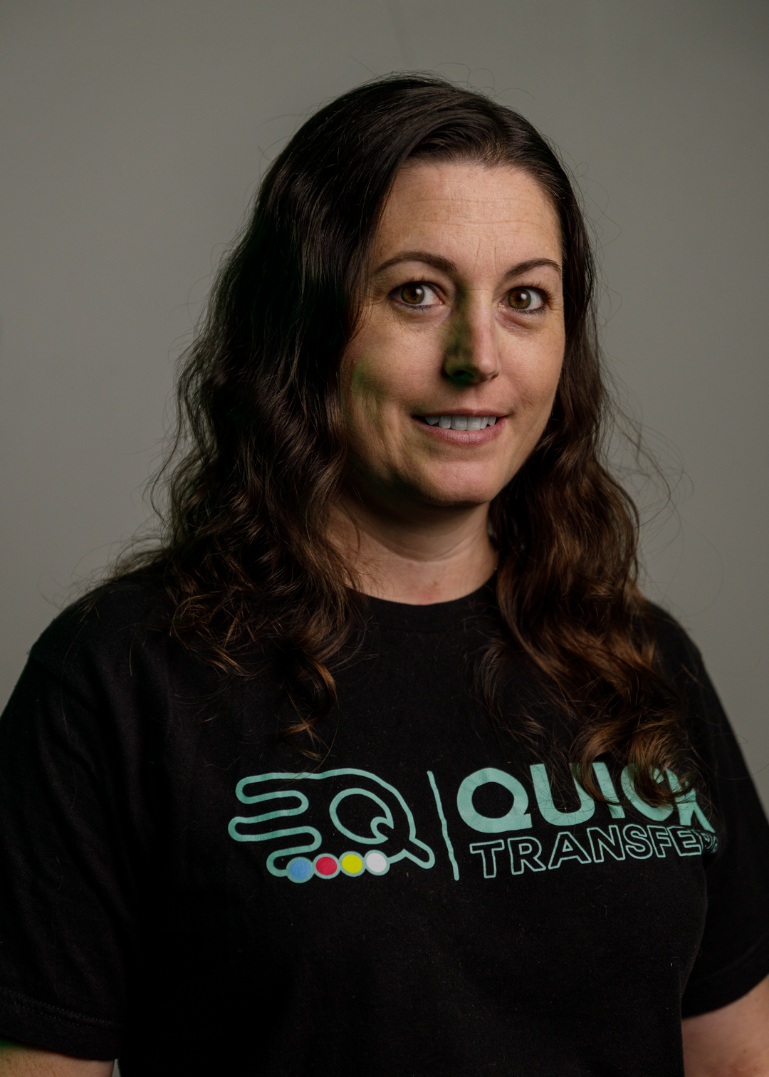 A person with long wavy hair smiles slightly, wearing a black T-shirt with the "Quick Transfer" logo, depicting a stylized bird. The background is plain and neutral, creating a professional portrait setting.