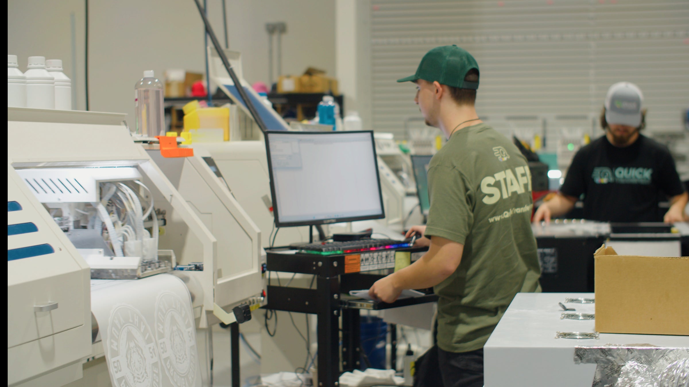 Two workers wearing green shirts and caps operate machines in a factory. One stands at a computer workstation, while the other, in the background, manages equipment. Shelving and materials are visible in the industrial setting.