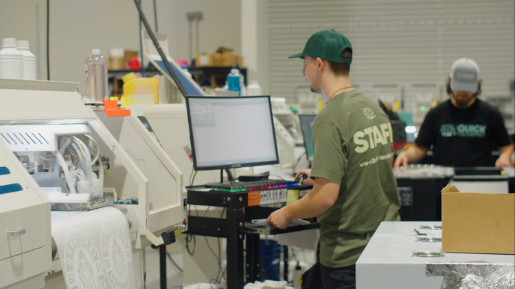 Two workers wearing green shirts and caps operate machines in a factory. One stands at a computer workstation, while the other, in the background, manages equipment. Shelving and materials are visible in the industrial setting.